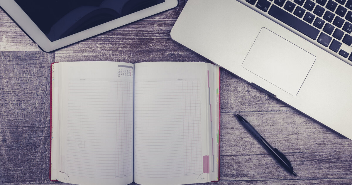 Flat-lay of an open notebook with a pen beside a laptop and tablet, representing editorial planning and restructuring content for GEO